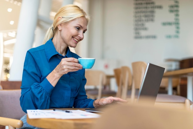 Vista lateral de la mujer de negocios mayor feliz tomando una taza de café y trabajando en la computadora portátil