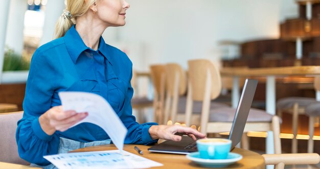 Vista lateral de la mujer de negocios mayor que trabaja en la computadora portátil mientras toma una taza de café