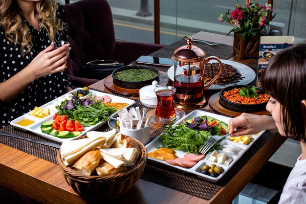 Vista lateral de una mujer con un niño desayunando una mesa servida huevos revueltos con hierbas con aperitivos vegetales panqueques pan y té