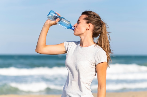 Vista lateral de la mujer que se mantiene hidratada durante el entrenamiento en la playa