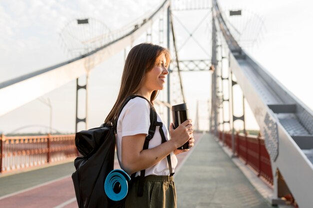 Vista lateral de la mujer que viaja feliz con mochila en puente sosteniendo termo