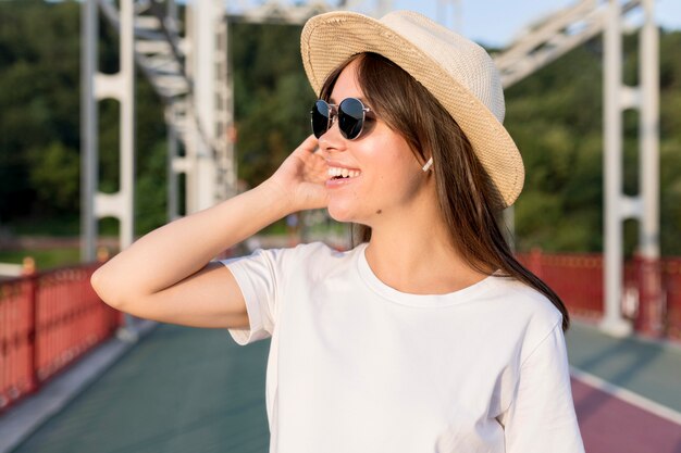 Vista lateral de la mujer que viaja sonriente en el puente con sombrero y gafas de sol