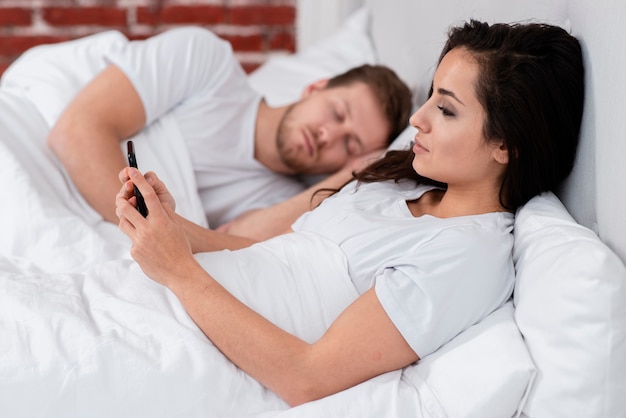 Vista lateral mujer revisando su teléfono junto al novio dormido