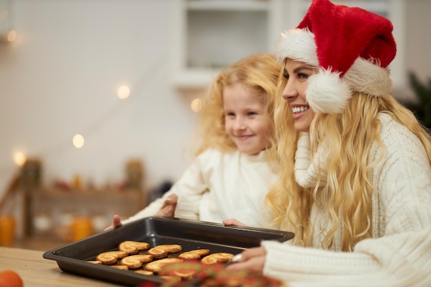 Foto gratuita vista lateral de una mujer rubia con sombrero de navidad horneando galletas con un niño pequeño mirando hacia adelante