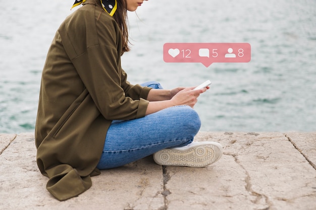 Foto gratuita vista lateral de la mujer sentada en el muelle con aplicación de redes sociales en el teléfono móvil