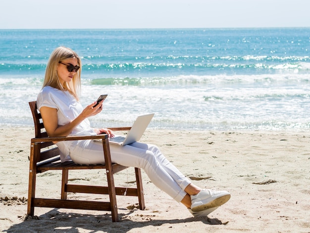 Vista lateral de la mujer en la silla de playa con computadora portátil y teléfono inteligente