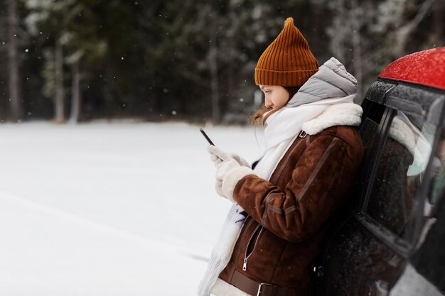 Vista lateral de la mujer con smartphone junto al coche durante un viaje por carretera de invierno