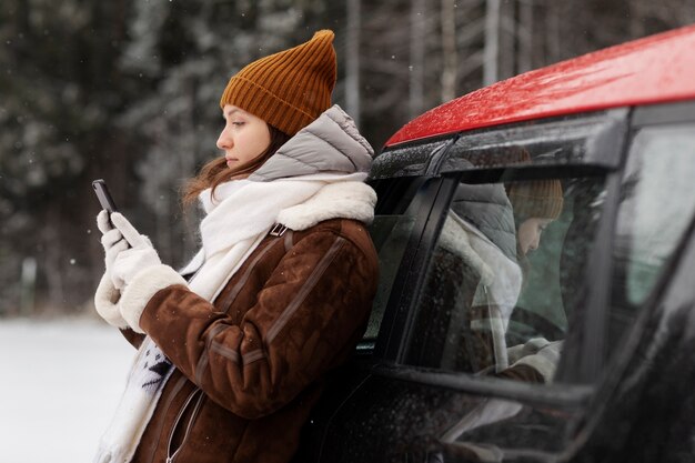 Vista lateral de la mujer con smartphone junto al coche durante un viaje por carretera de invierno