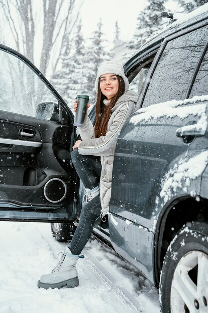 Vista lateral de la mujer sonriente disfrutando de la nieve durante un viaje por carretera y tomando una bebida caliente