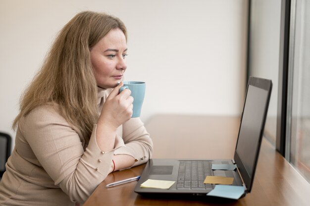 Vista lateral de la mujer sosteniendo la taza y trabajando en la computadora portátil en la oficina