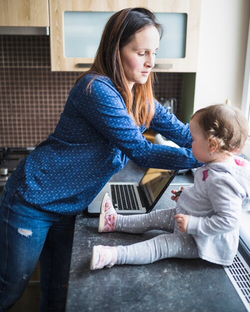 Vista lateral de la mujer y su hija con la computadora portátil en la encimera de la cocina