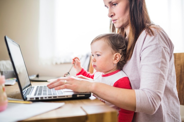 Vista lateral de una mujer con su niño usando la computadora portátil en el escritorio de madera
