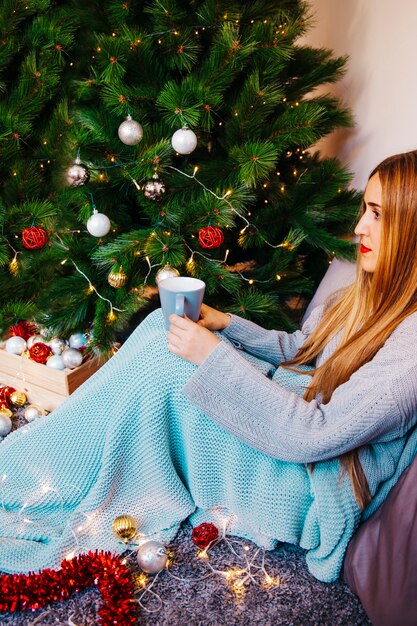 Vista lateral de mujer sujetando taza enfrente de árbol de navidad