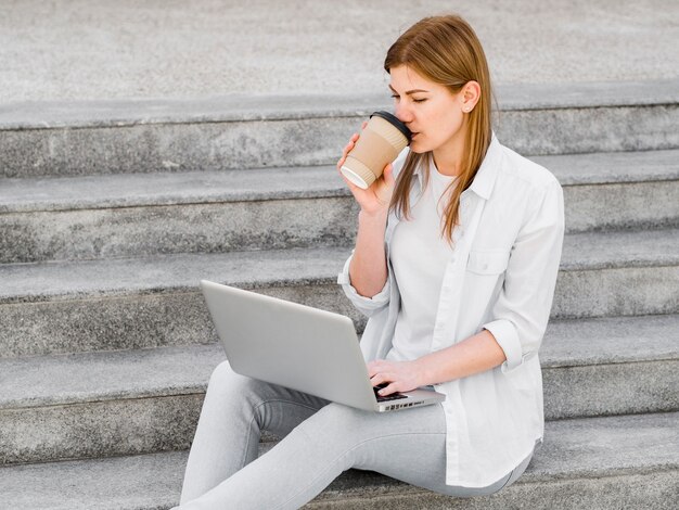 Vista lateral de la mujer tomando café en pasos mientras trabajaba en la computadora portátil