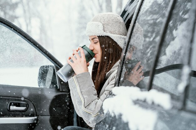 Vista lateral de la mujer tomar una bebida caliente y disfrutar de la nieve durante un viaje por carretera