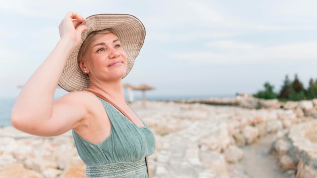 Vista lateral de la mujer turista senior posando con sombrero de playa