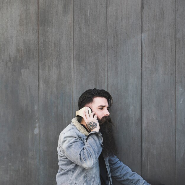 Vista lateral de una música que escucha del hombre joven en el auricular contra la pared de madera gris