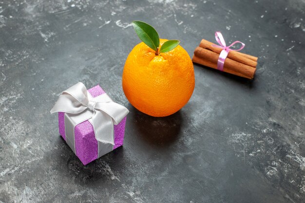 Vista lateral de naranja fresca orgánica con tallo y hoja cerca de un regalo y limones de canela sobre fondo oscuro