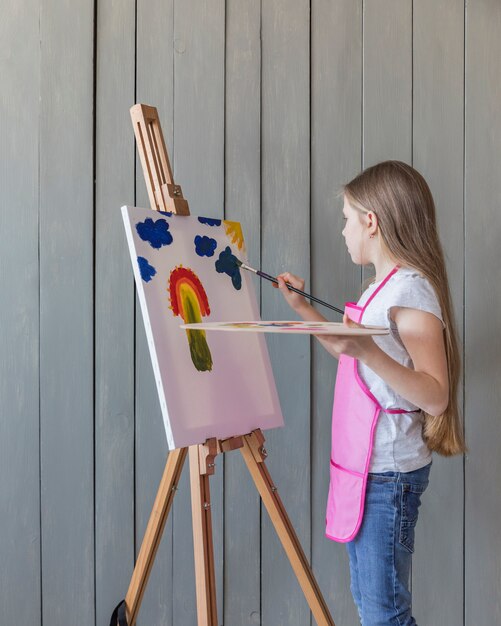 Vista lateral de una niña dibujando con pincel sobre caballete contra un tablón de madera