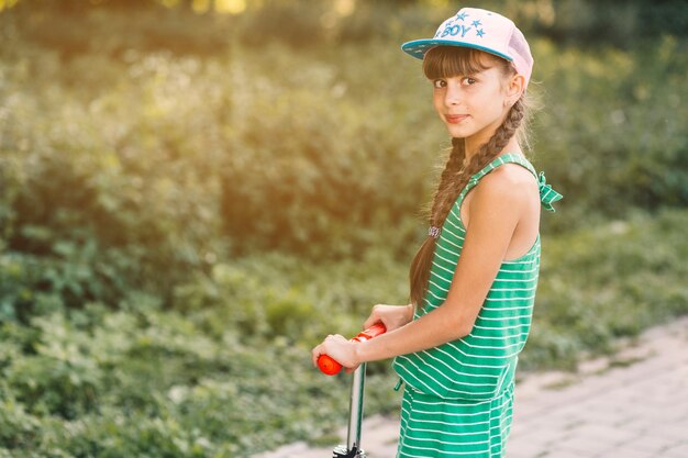 Vista lateral de una niña con gorra de pie en patinete scooter