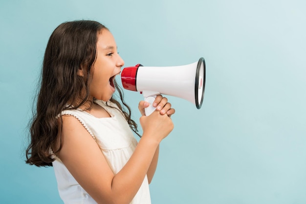 Vista lateral de una niña gritando en un megáfono mientras está de pie contra un fondo liso