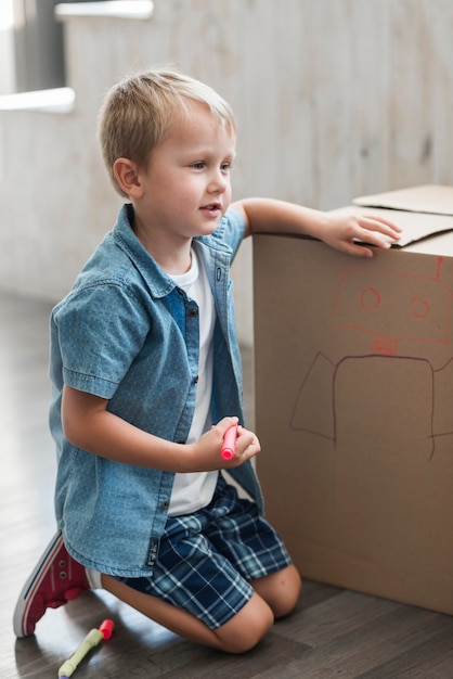 Vista lateral de un niño dibujando con un marcador en una caja de cartón