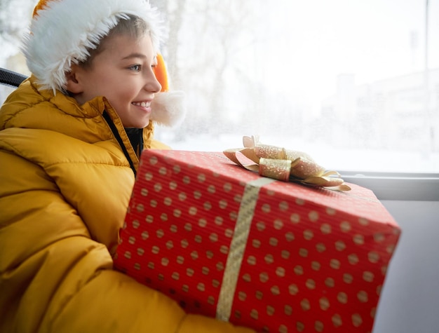 Foto gratuita vista lateral de un niño feliz con sombrero naranja de papá noel y un regalo en papel rojo de lunares mirando hacia el futuro