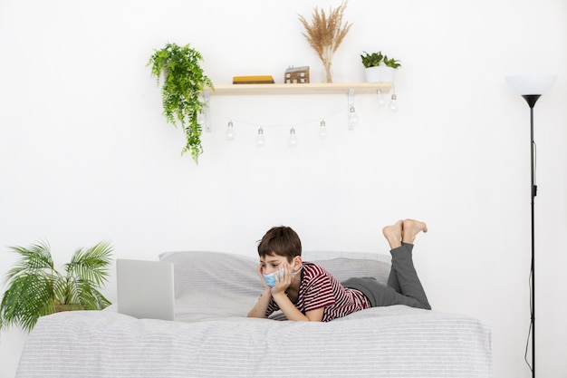 Vista lateral del niño con máscara médica viendo algo en la computadora portátil en la cama