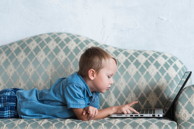 Vista lateral de un niño rubio acostado en el sofá usando laptop