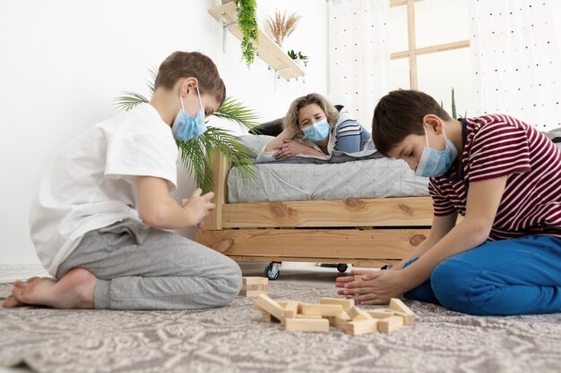 Vista lateral de niños en casa usando máscaras médicas y jugando jenga