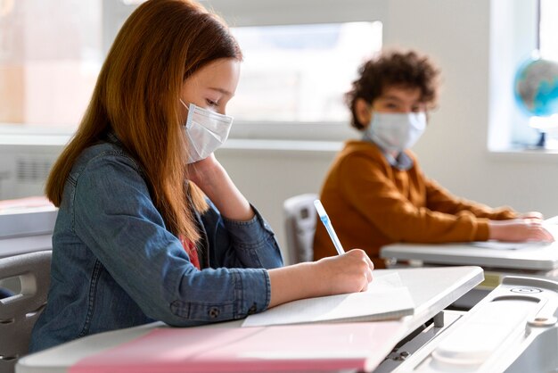 Vista lateral de los niños con máscaras médicas en el aula estudiando