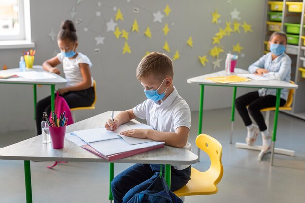 Vista lateral de los niños tomando notas en clase mientras usan máscaras médicas