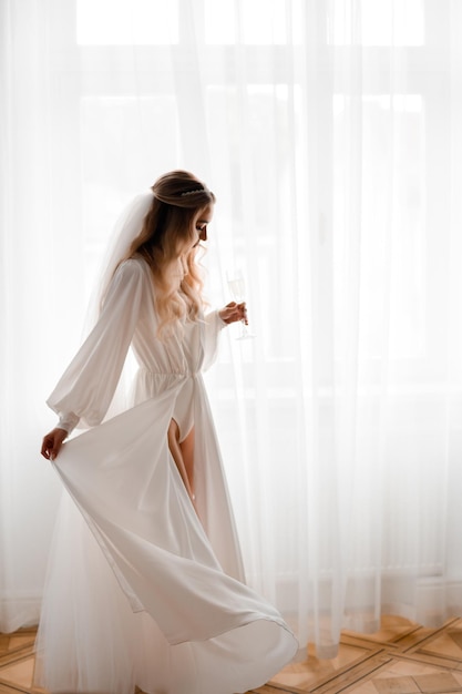 Foto gratuita vista lateral de la novia bonita con peinado largo y rizado y velo vestido con lencería blanca y peignoir largo sosteniendo un vaso con bebida y girando cerca de la ventana en la sala de luz durante la mañana de la boda