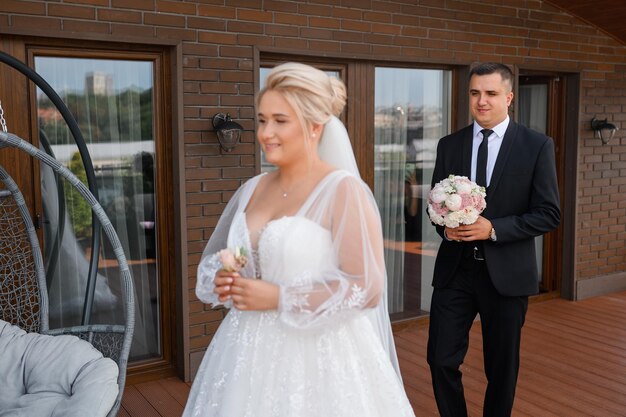 Vista lateral del novio feliz con traje de moda sosteniendo un ramo de novia y entrando en la terraza para encontrarse con la novia que le da la espalda con un ojal de boda y esperando la primera cita