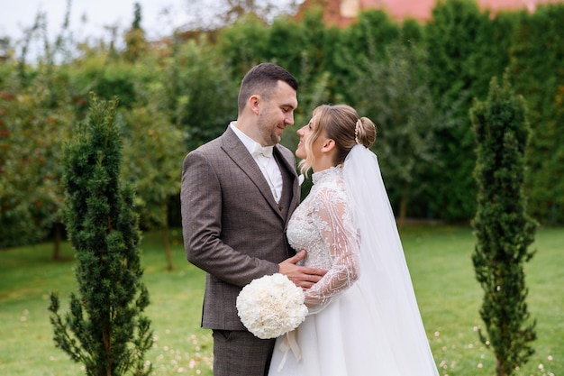 Vista lateral del novio y la novia felices vestidos con traje y vestido de novia parados en el jardín cerca de hermosas plantas sonriendo y mirándose el uno al otro el día de la boda