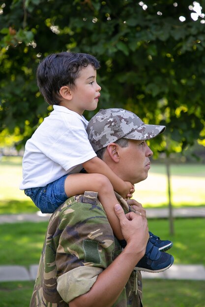 Vista lateral del padre sosteniendo a su hijo en el cuello y caminando en el parque de la ciudad. Hijo caucásico sentado en el cuello de papá en uniforme militar, abrazándolo y mirando hacia adelante. Concepto de paternidad y regreso a casa