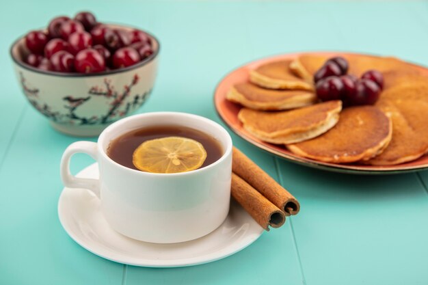 Vista lateral de panqueques con cerezas en plato y taza de té con rodaja de limón y canela en platillo y tazón de cerezas sobre fondo azul.