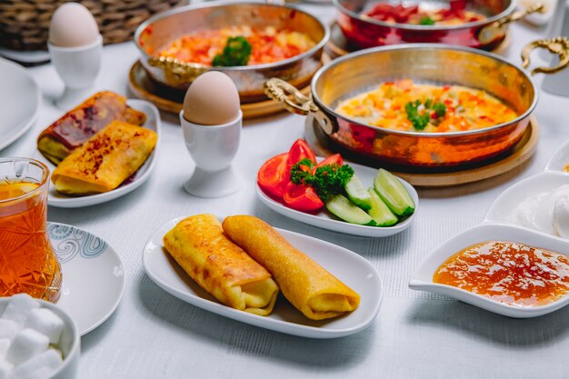 Vista lateral panqueques enrollados con huevos cocidos, tomates, pepinos y miel sobre la mesa servida el desayuno