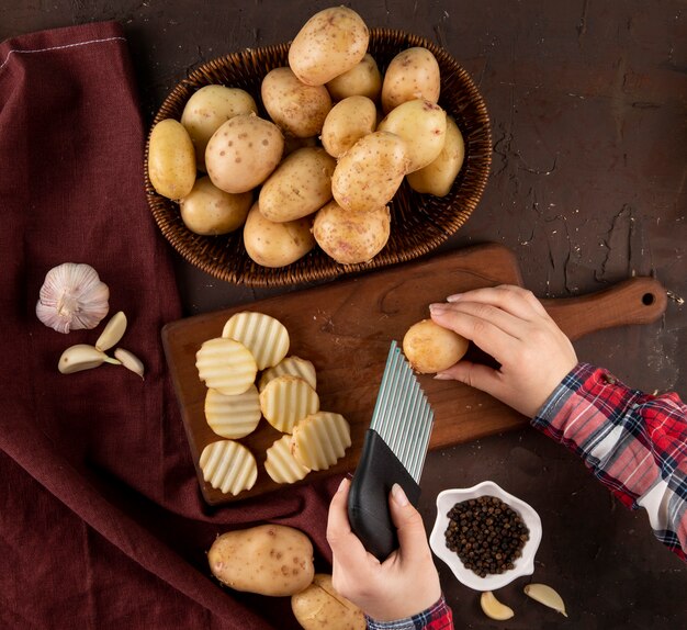 Vista lateral de papas en una canasta una mujer rebana papas en un tablero con pimienta negra