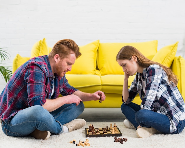 Vista lateral de una pareja joven sentada en una alfombra blanca jugando al juego de ajedrez
