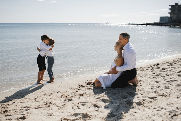 Vista lateral de la pareja que está sentada en la playa de arena cerca del mar y mirando a los dos pequeños hijos que se abrazan