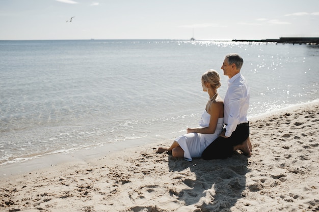Vista lateral de la pareja que está sentada en la playa de arena cerca del mar y mirando el impresionante paisaje