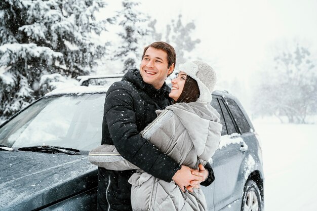 Vista lateral de la pareja sonriente abrazándose en la nieve durante un viaje por carretera