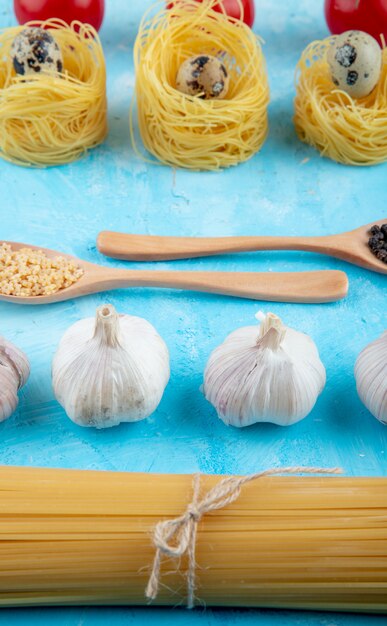 Vista lateral de pasta cruda de diferentes formas y tipos como espagueti pasta amarilla nido con pequeños huevos de codorniz cuchara de madera con pasta en forma de estrella y ajo en azul