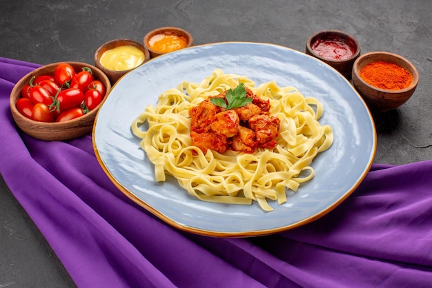 Vista lateral de la pasta y las salsas plato azul de pasta, hierbas, carne y salsa entre tomates y salsas de colores en tazones sobre el mantel púrpura