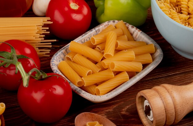 Vista lateral de pasta ziti en un tazón con spaghetti y rotini tipos en un tazón de sal, tomate, ajo, pimienta en la mesa de madera