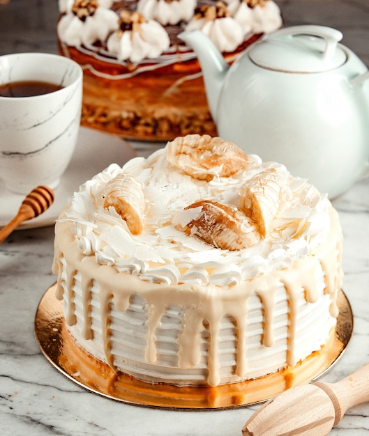 Vista lateral del pastel blanco decorado con crema batida de chocolate blanco derretido y plátanos sobre la mesa