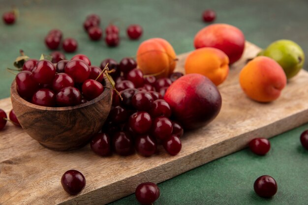 Vista lateral del patrón de frutas como albaricoques, melocotones, cerezas de pera con tazón de cereza en la tabla de cortar y sobre fondo verde