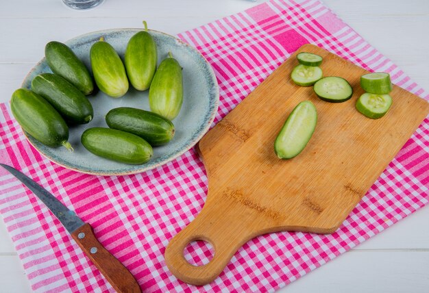 Vista lateral de pepinos cortados y rebanados en tabla de cortar con enteros en plato y cuchillo en tela escocesa y mesa de madera