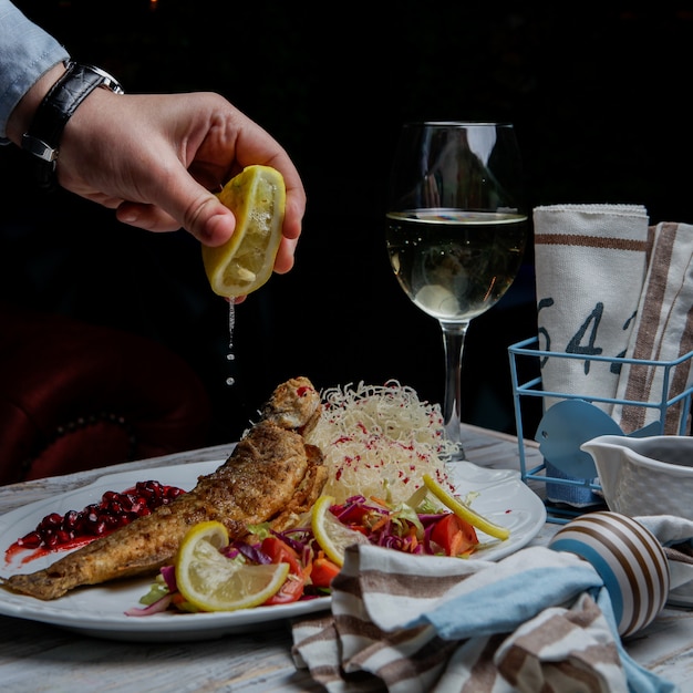 Vista lateral pescado frito con copa de vino y limón y mano humana en plato blanco